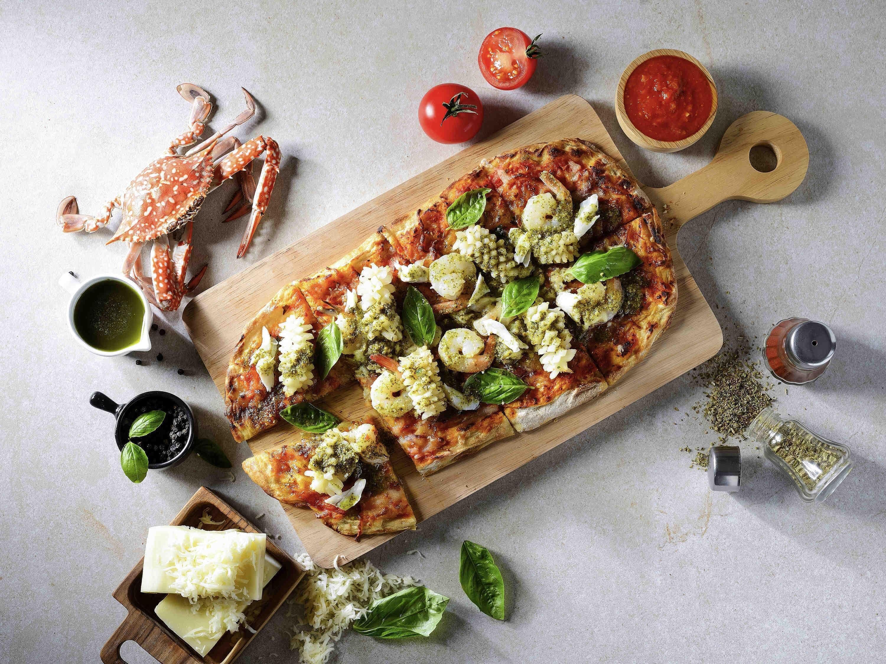 Une pizza garnie de divers ingrédients, présentée sur une planche en bois, entourée d’autres ingrédients et d’ustensiles.
