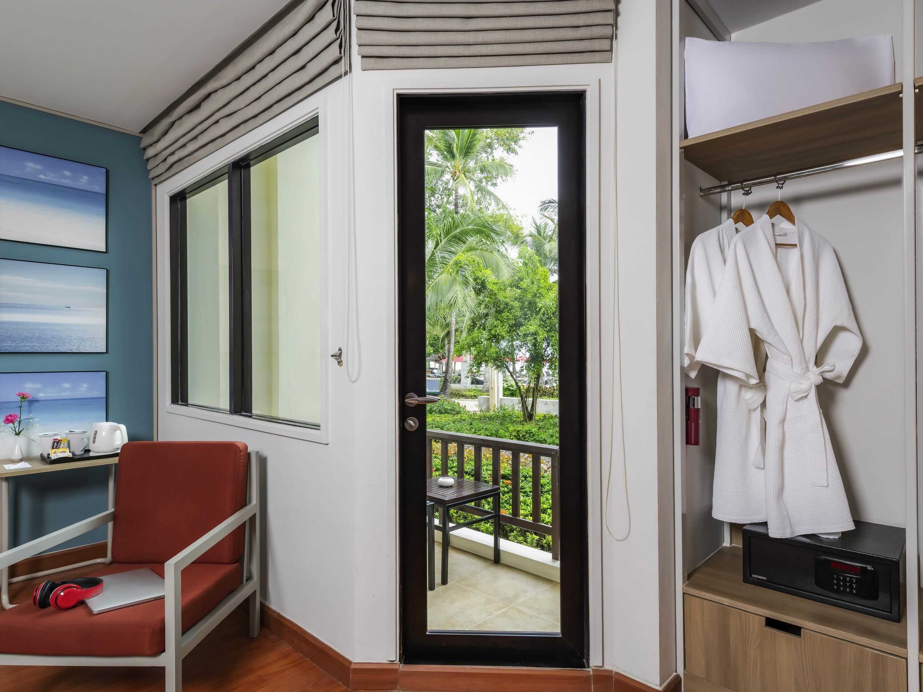 A hotel room with a large window, a red chair, a table with a laptop and headphones, a robe hanging on a rack, and a microwave on a wooden cabinet.