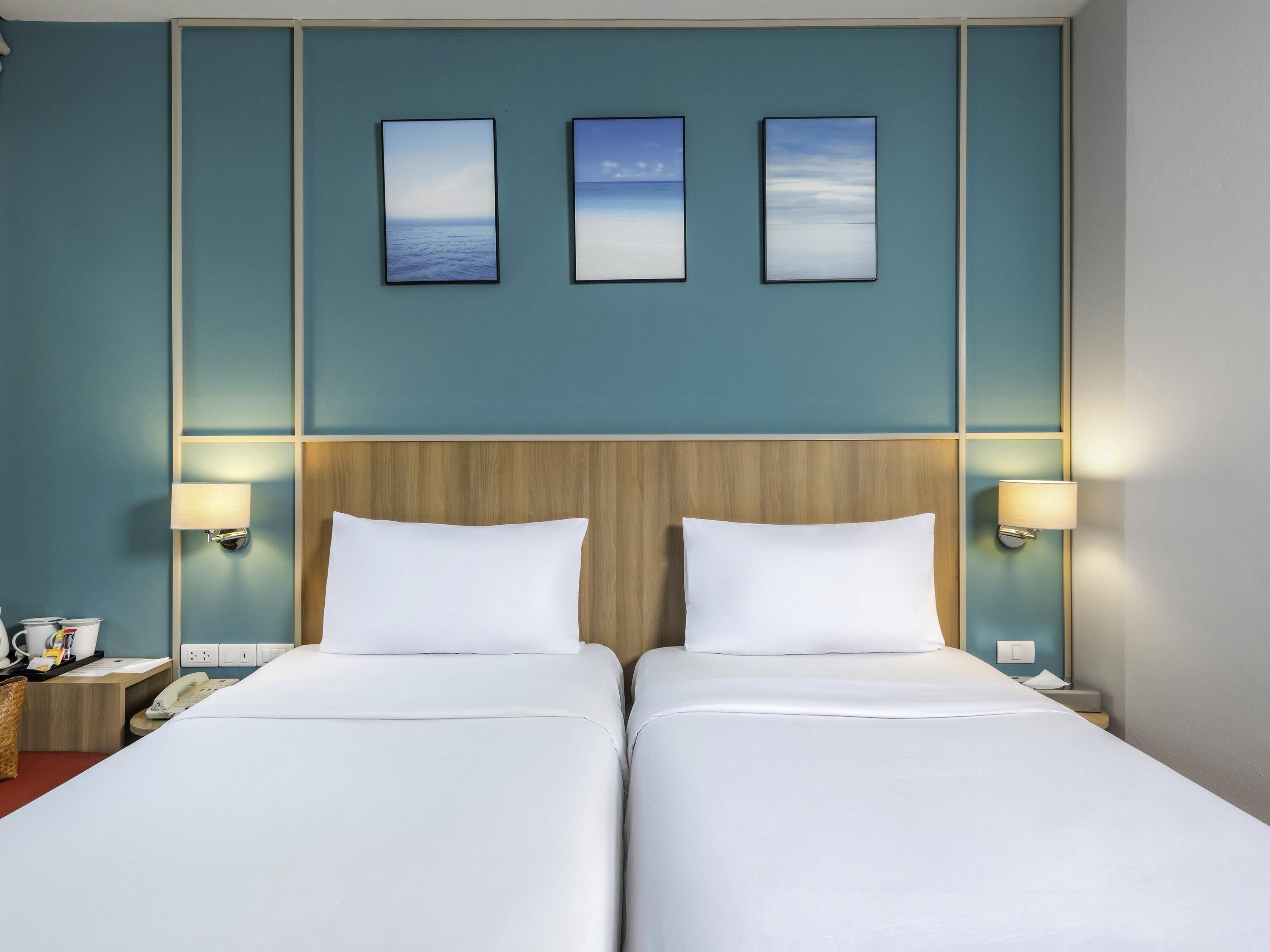 A hotel room with two beds, a wooden headboard, wall-mounted lamps, and three framed pictures above the beds.