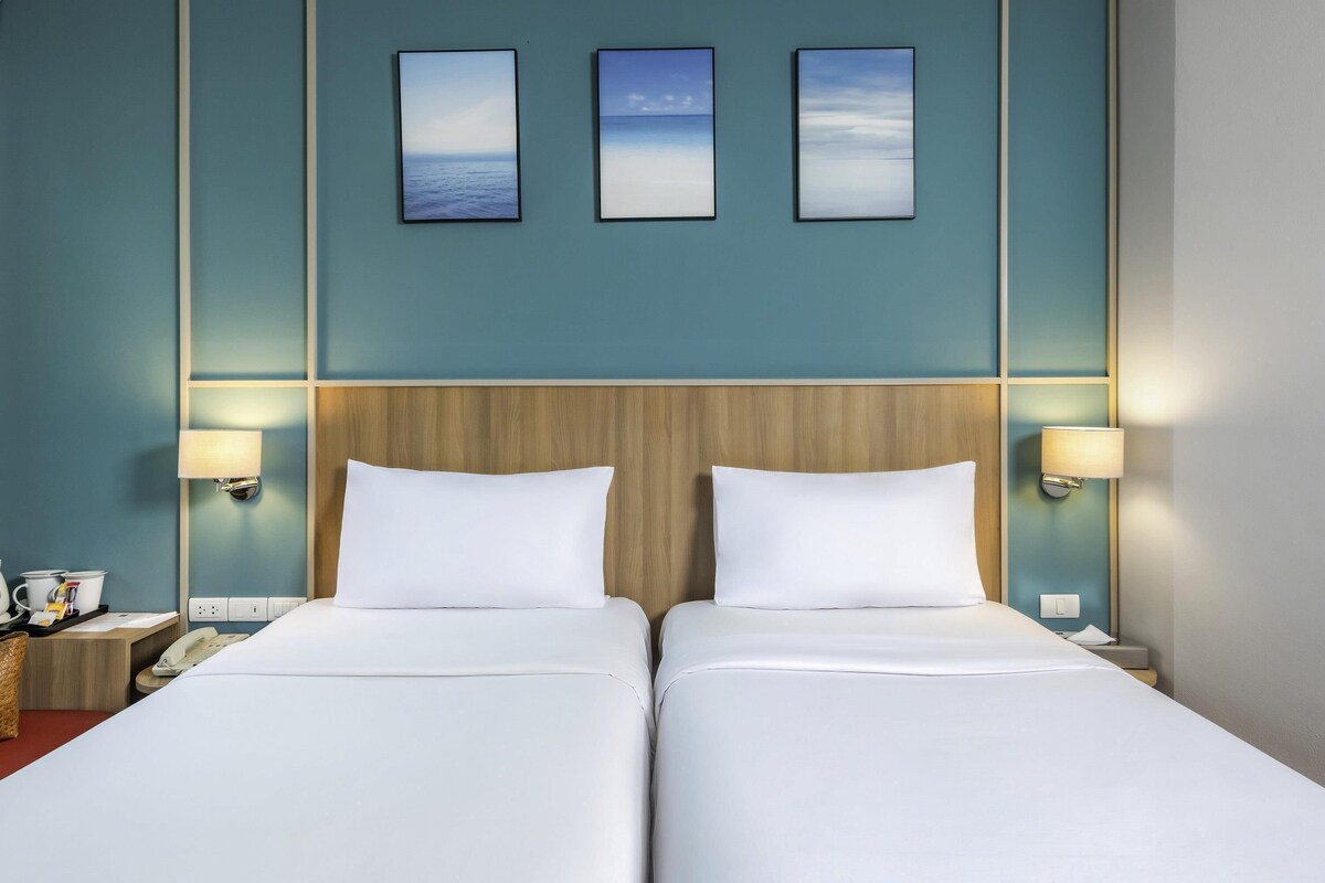 Habitación de hotel con dos camas, cabecero de madera, lámparas de pared y tres cuadros enmarcados sobre las camas.