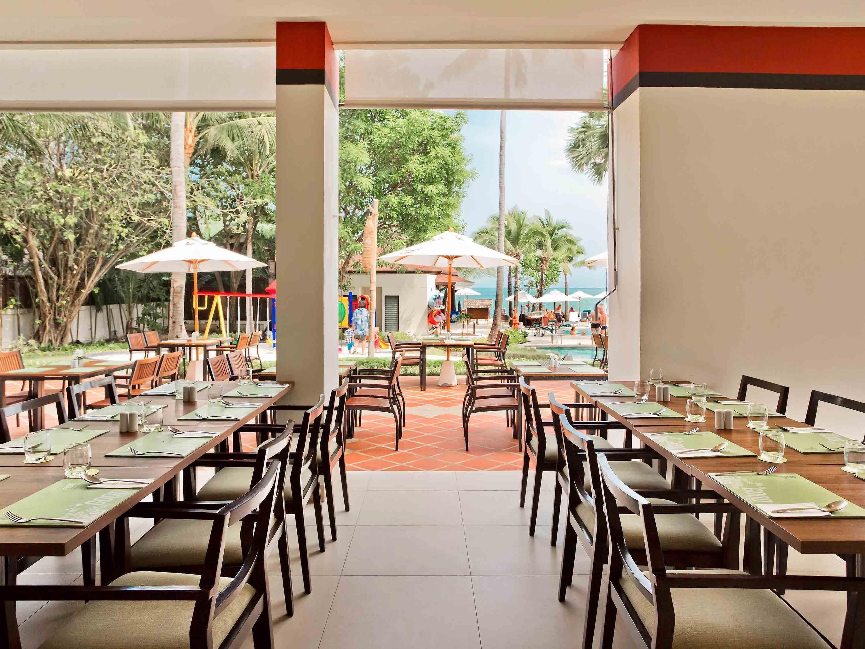 En restaurant med udendørs siddepladser, borde med grønne servietter og parasoller.