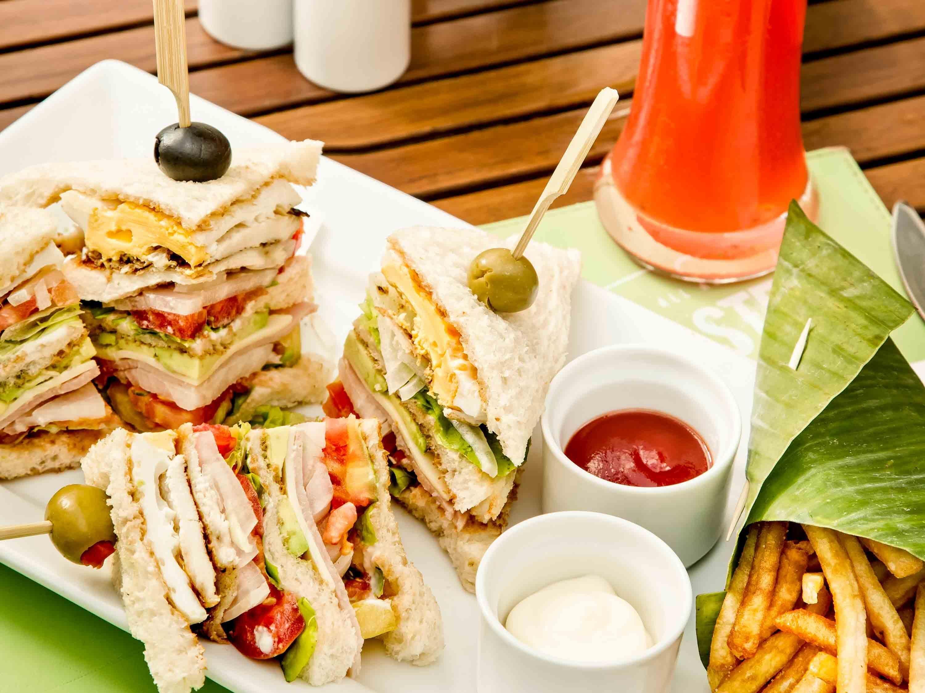 A sandwich cut into halves with a toothpick, accompanied by a small bowl of ketchup and a container of mayonnaise, and a side of French fries.