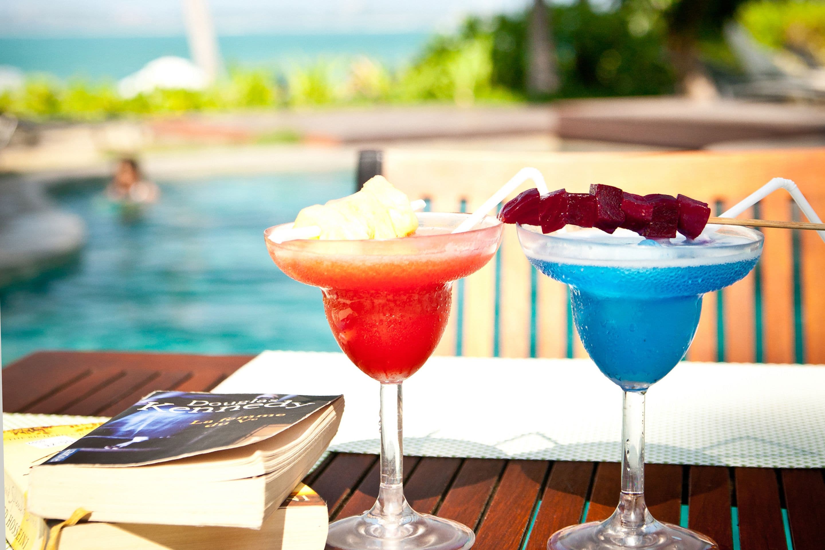 Twee cocktails op een houten tafel, met een boek en een zwembad op de achtergrond.