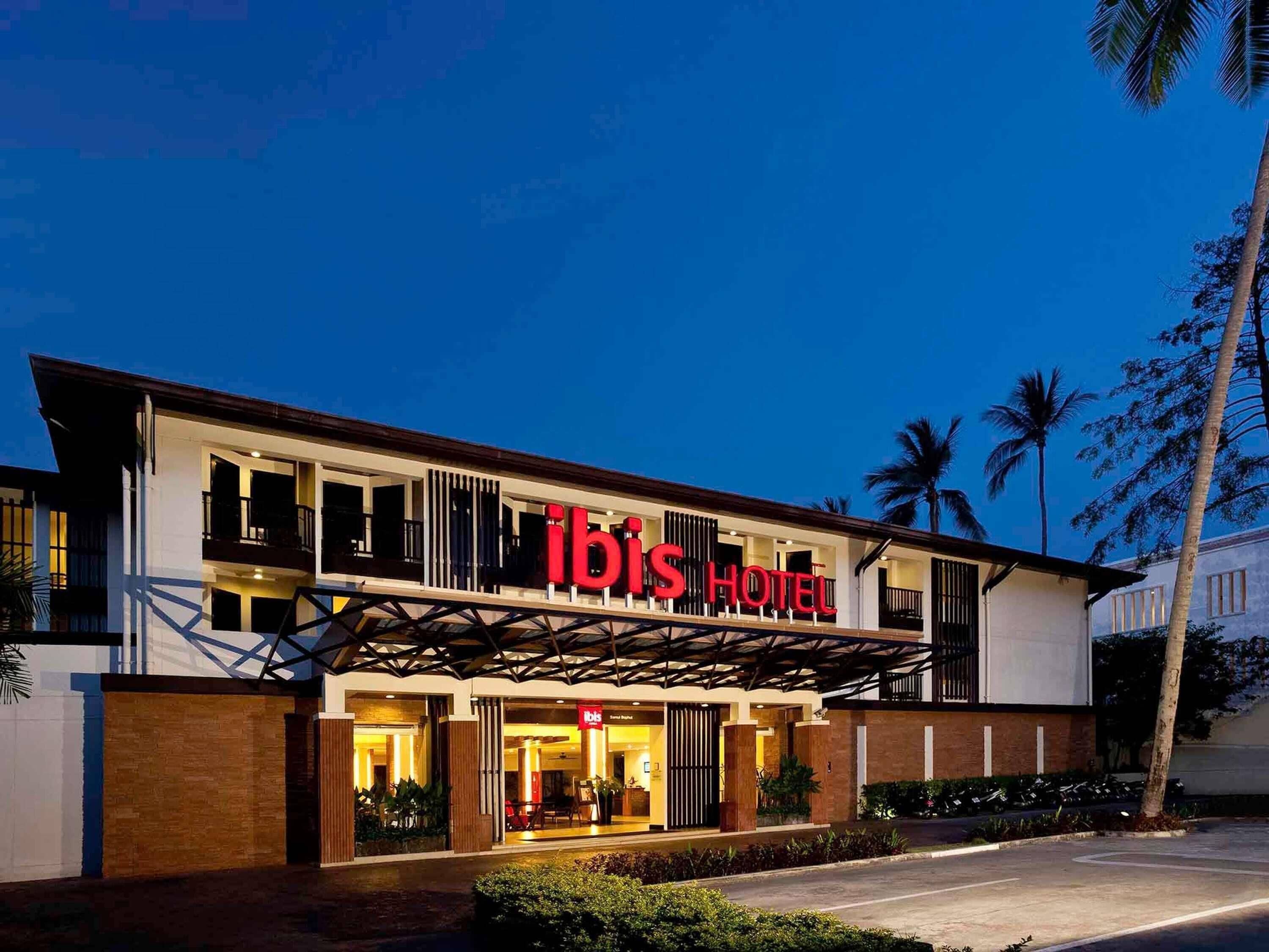 A modern hotel with a prominent 'ibis Hotel' sign, a covered entrance, and a landscaped front yard.