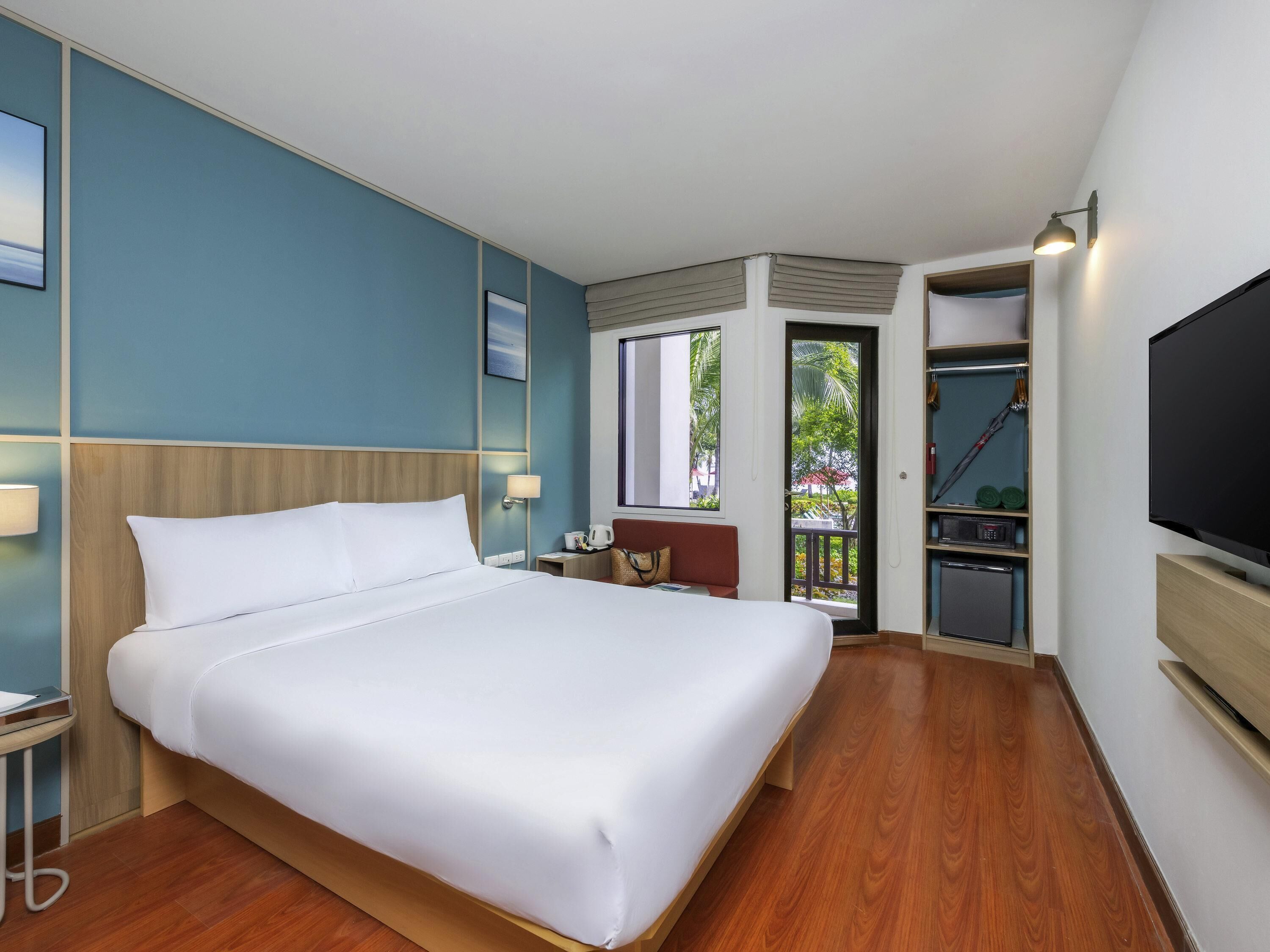 Habitación de hotel con una cama grande, televisión, escritorio con silla, pequeña mesa con lámpara y puerta que da a un balcón.