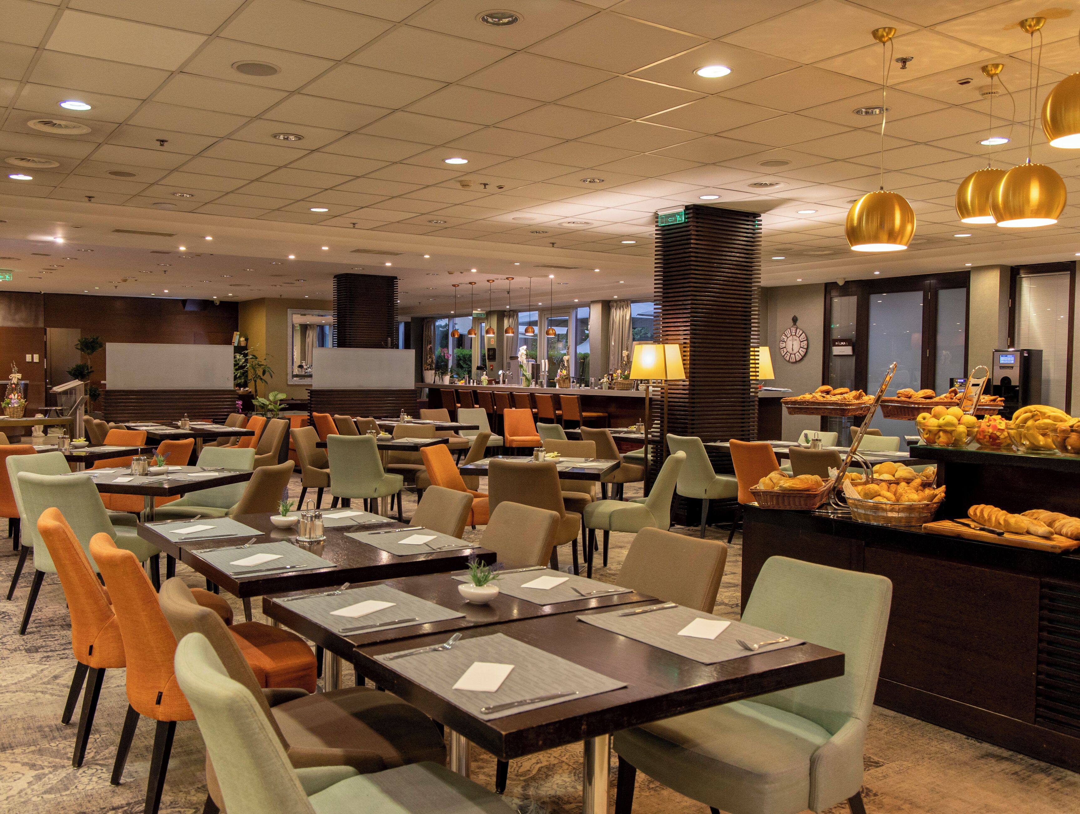 Espaço de refeições de hotel com várias mesas e cadeiras, um buffet com diversos alimentos e uma tela grande para apresentações.