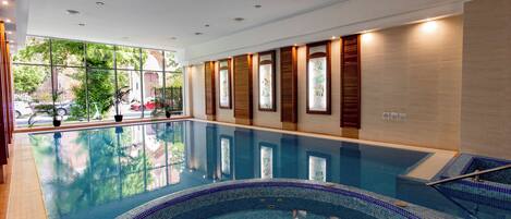 A large indoor swimming pool with clear glass walls, a tiled edge, and a wooden deck area.