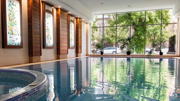 A large indoor swimming pool with clear water, wooden paneling, and large windows offering a view of the outside. - Lions Garden Hotel (Budapest)