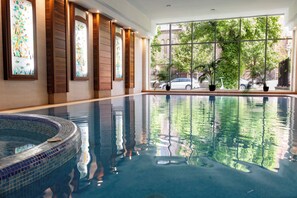A large indoor swimming pool with clear water, wooden paneling, and large windows offering a view of the outside. - Lions Garden Hotel (Budapest)