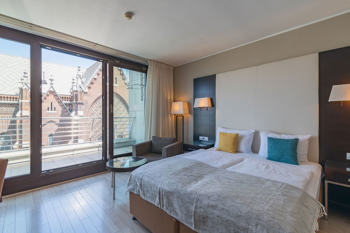 A hotel room with a large bed, a chair, a small table, and a view of buildings through a large window.
