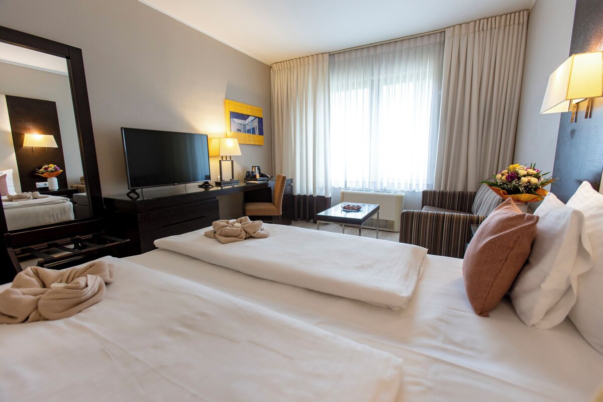 A hotel room with two beds, a desk, a chair, a television, and a large window with curtains.