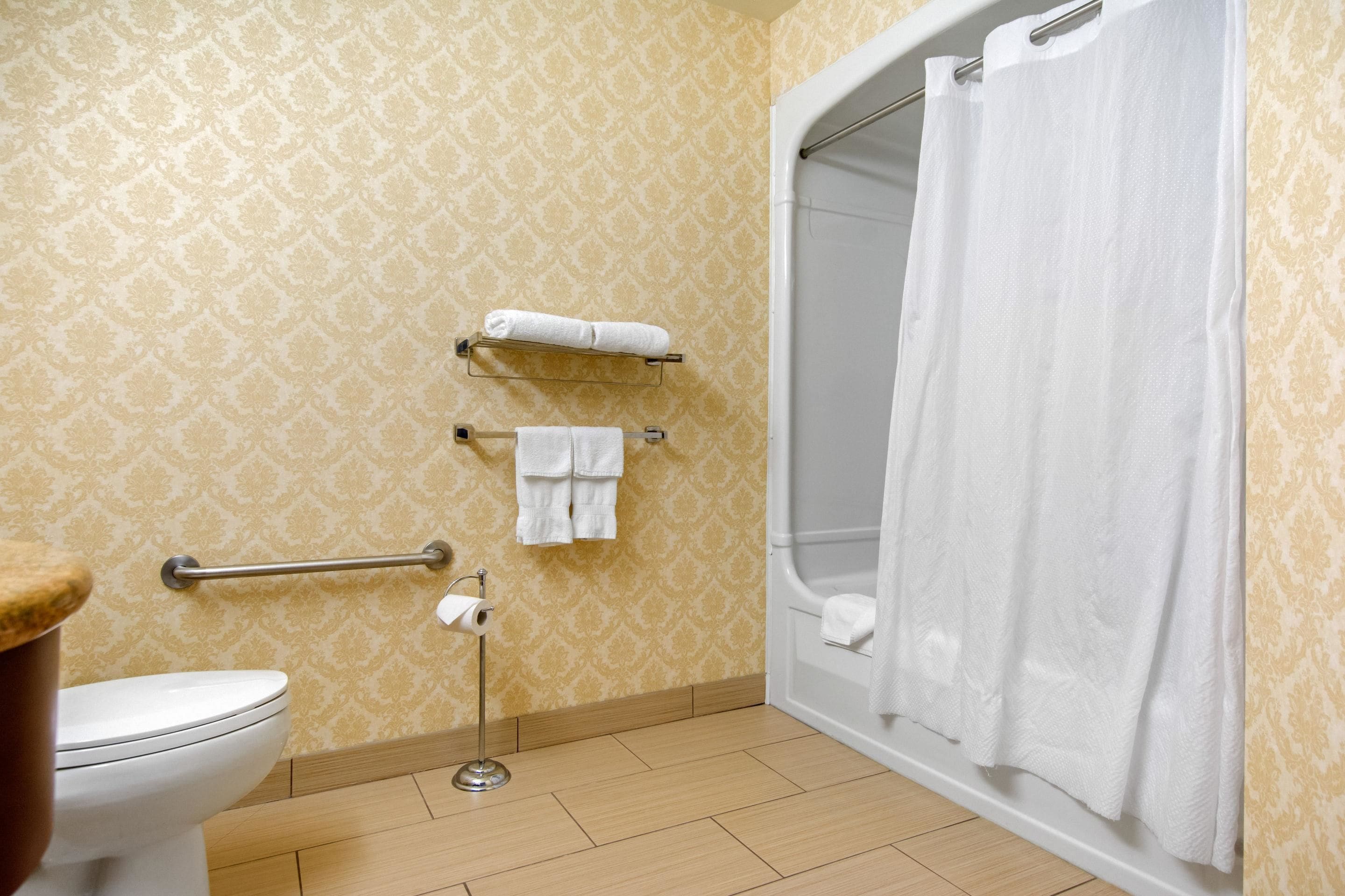 A bathroom with a toilet, a handrail, a towel rack with towels, a shower curtain, and a patterned wall.