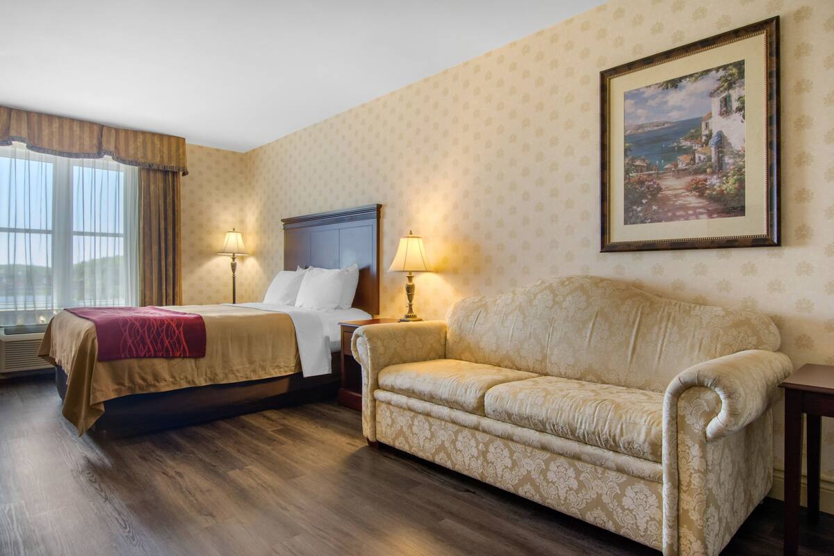A hotel room with a bed, a sofa, two bedside lamps, a window with curtains, and a framed picture on the wall.