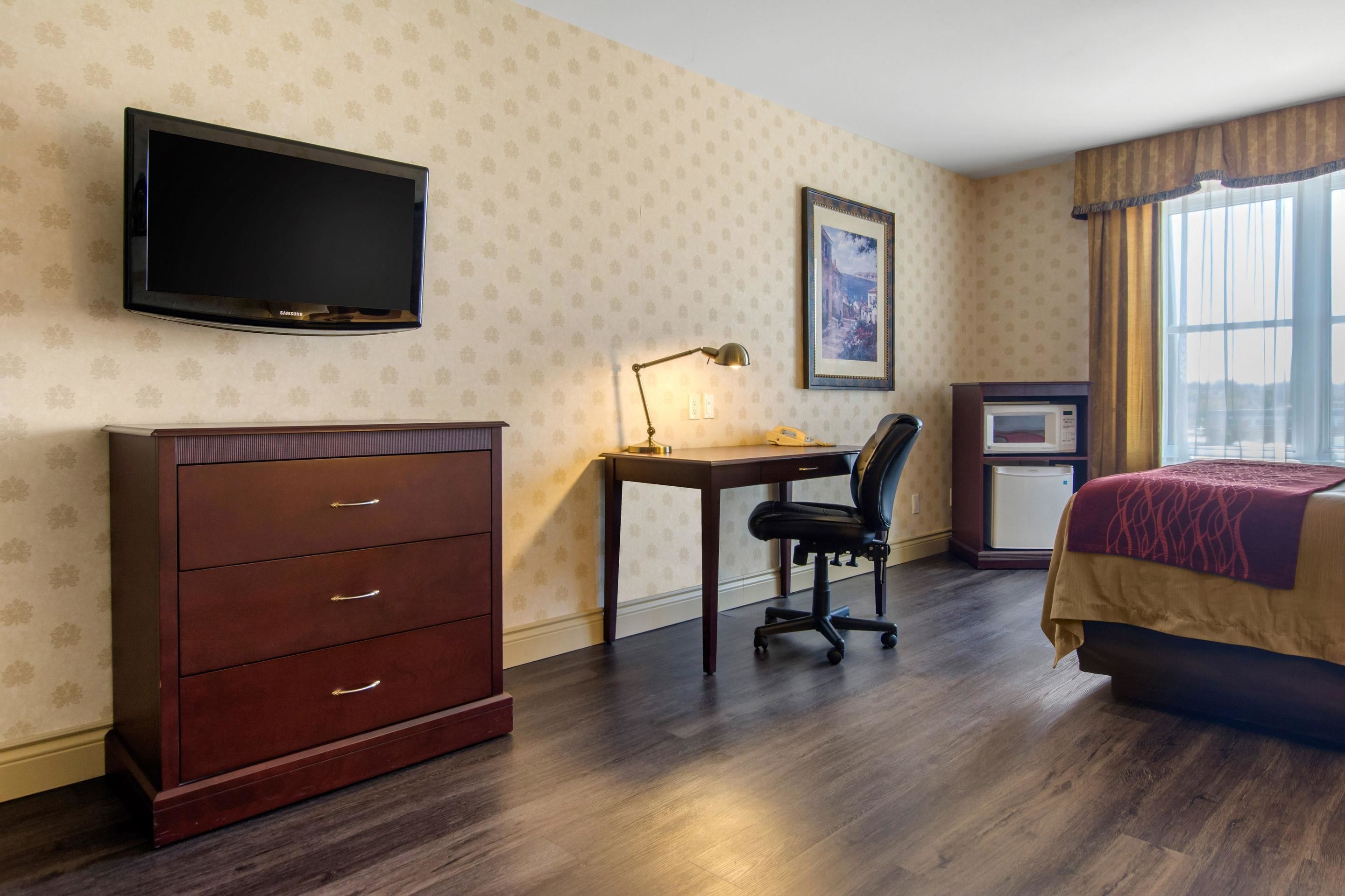 Une chambre d’hôtel équipée d’une télévision à écran plat, d’un bureau avec une lampe, d’une chaise, d’un lit avec une couverture rouge et d’un petit réfrigérateur.