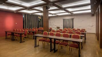 A conference room with rows of tables and chairs, wooden floors, and a high ceiling with exposed beams and lighting fixtures.