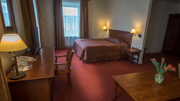 A hotel room with a bed, bedside tables, a desk, a chair, a lamp, and a vase with flowers. - Daugirdas Hotel (Kaunas)