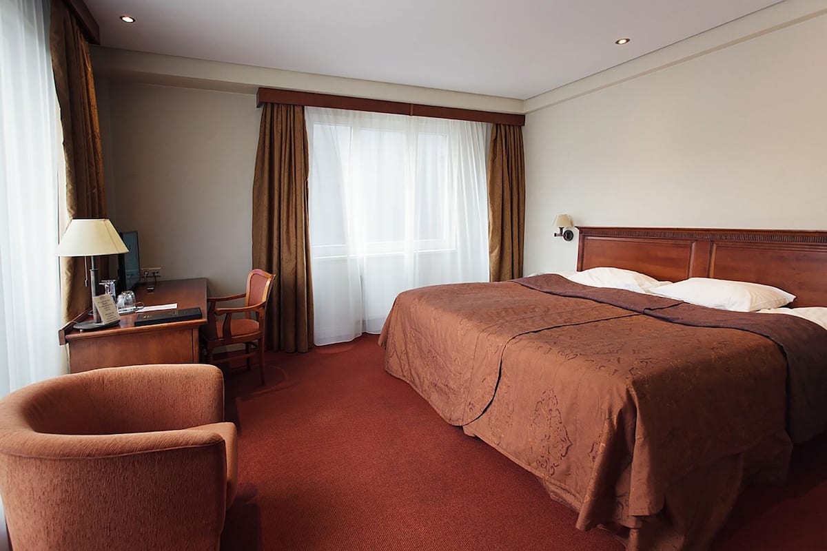 Ein Hotelzimmer mit einem großen Bett, einem Schreibtisch mit Lampe, einem Sessel und einem Fenster mit Vorhängen.