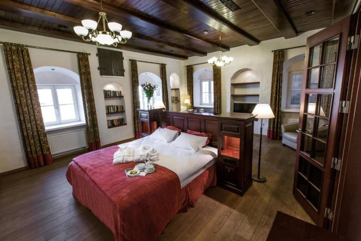 Ein großzügiges Schlafzimmer mit einem großen Bett, Holzboden und einem Kronleuchter. Ein Nachttisch mit Lampe und eine kleine Regal mit Büchern sind vorhanden.