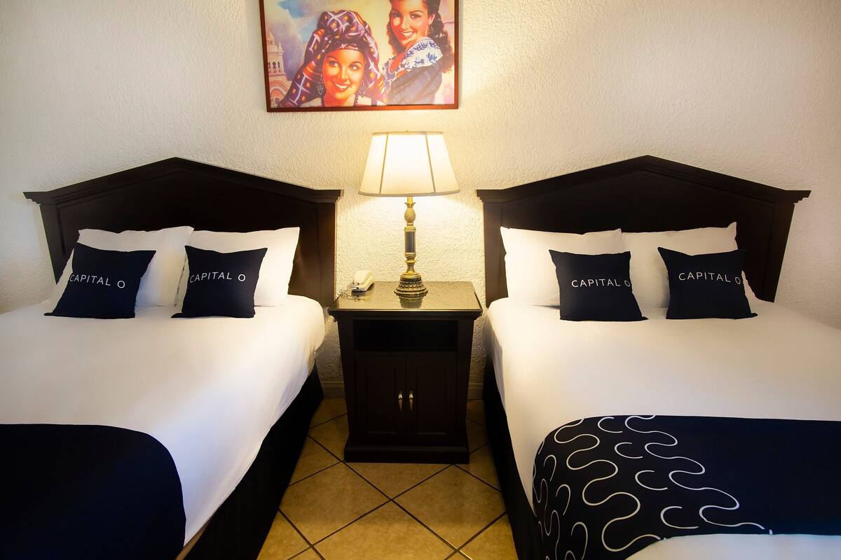 Quarto de hotel com duas camas, cada uma com cabeceira, roupa de cama branca e almofadas com a etiqueta “CAPITAL O”. Há uma mesa de cabeceira com uma luminária entre as camas, e um quadro na parede acima da mesa.