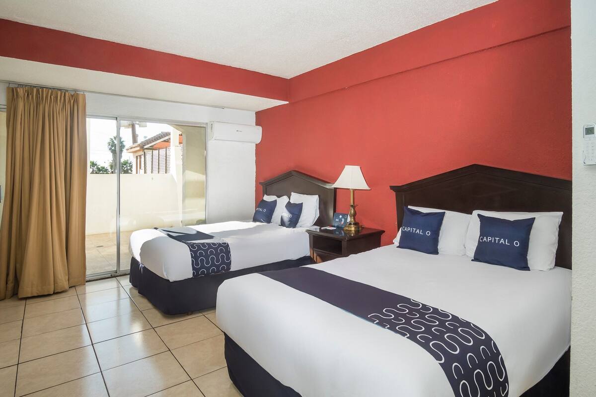 Quarto de hotel com duas camas, porta de correr de vidro para o exterior e parede vermelha.
