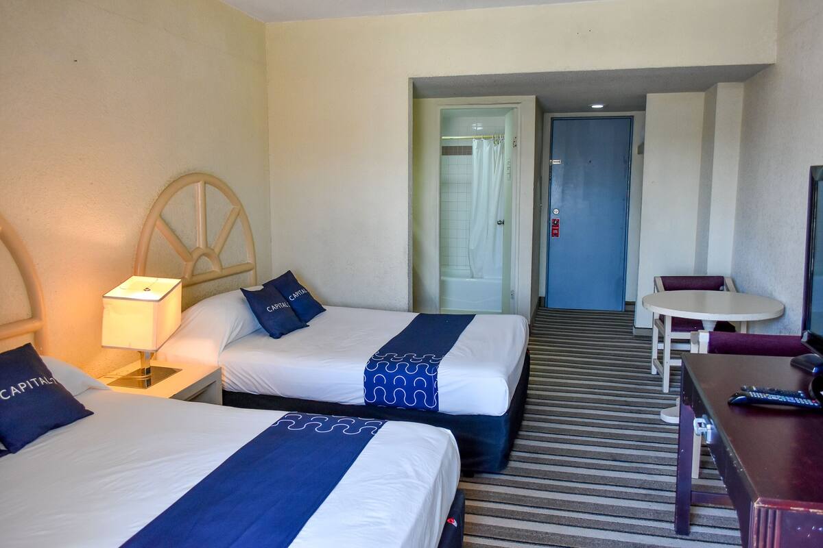 Quarto de hotel com duas camas, uma escrivaninha, uma cadeira e um banheiro visível por uma porta aberta.