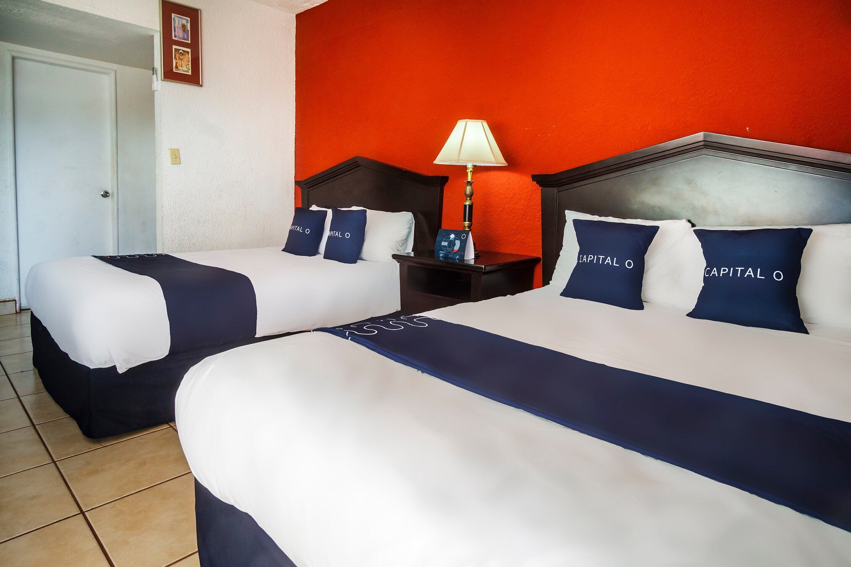 Two beds with matching navy blue and white bedding, a nightstand with a lamp, and a red wall.
