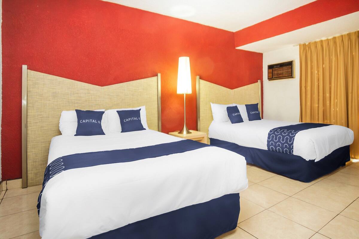 Quarto de hotel com duas camas, cada uma com cabeceira, mesa de cabeceira, abajur e janela com cortinas.