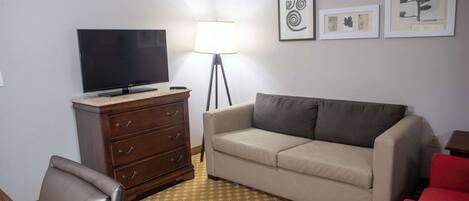 A room with a flat-screen TV on a wooden dresser, a beige sofa, a red chair, a patterned carpet, and framed artwork on the wall.