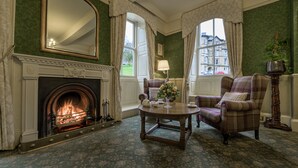 A room with a fireplace, a plaid armchair, a round table with a vase of flowers, and a view of buildings through the windows. - Old Hall Hotel (Buxton)