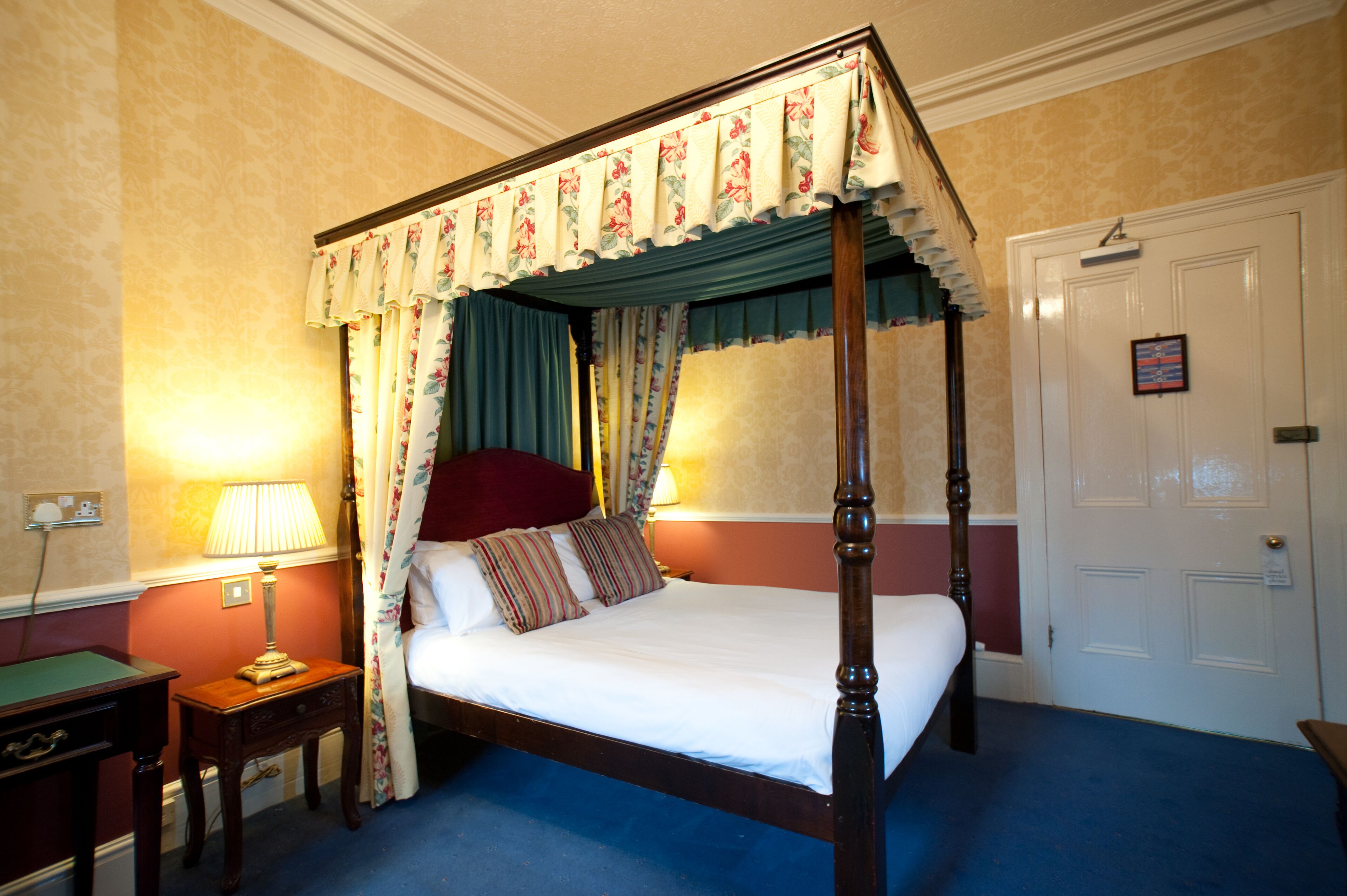 A four-poster bed with floral curtains, a bedside lamp, and a door with a picture frame.