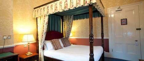 A four-poster bed with floral curtains, a bedside lamp, and a door with a picture frame.