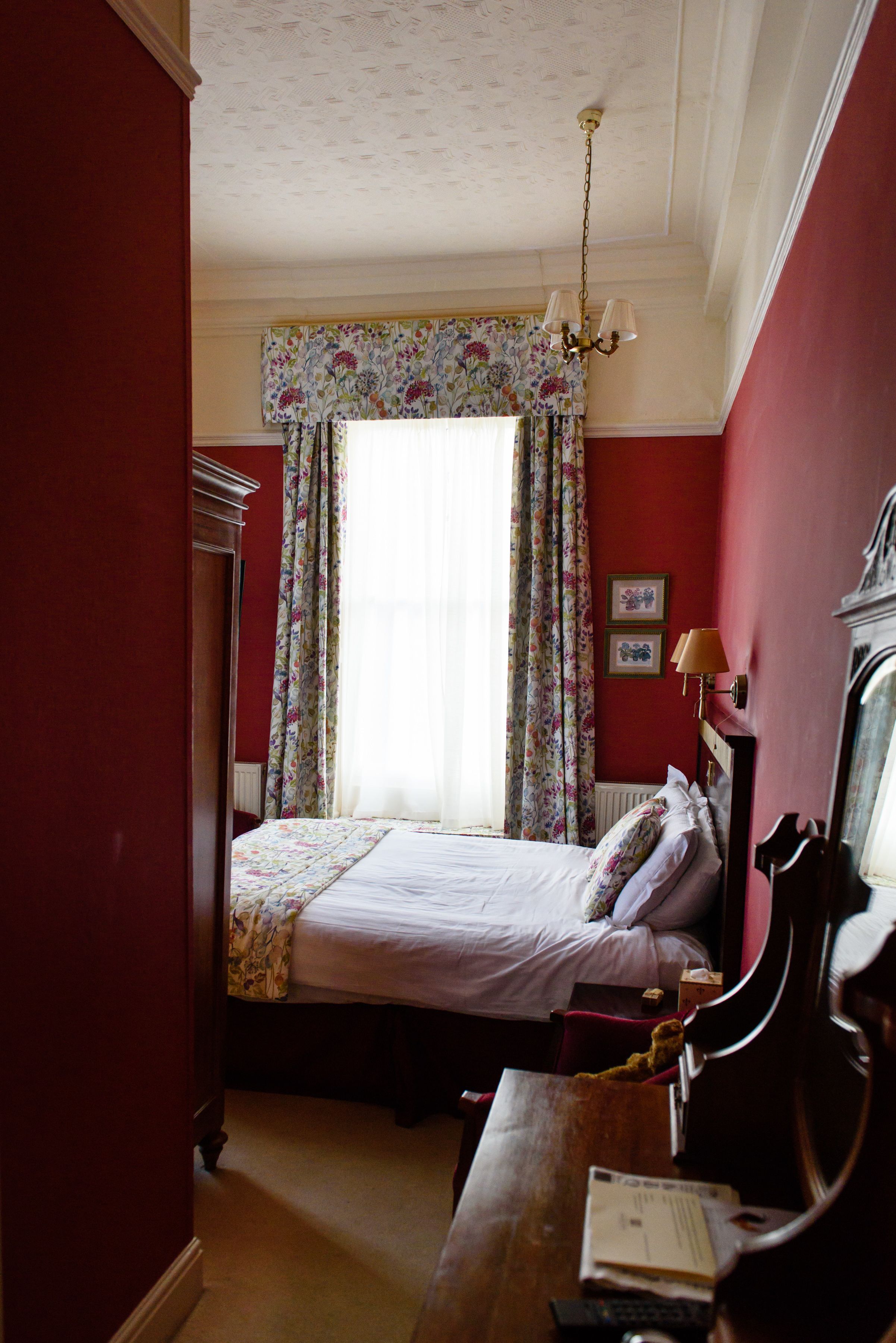 A bedroom with a bed, a desk, a chair, a dresser, a window with floral curtains, and a chandelier.