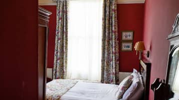A bedroom with a bed, a desk, a chair, a dresser, a window with floral curtains, and a chandelier.
