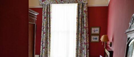 Ein Schlafzimmer mit Bett, Schreibtisch, Stuhl, Kommode, einem Fenster mit floralen Vorhängen und einer Chandelier.