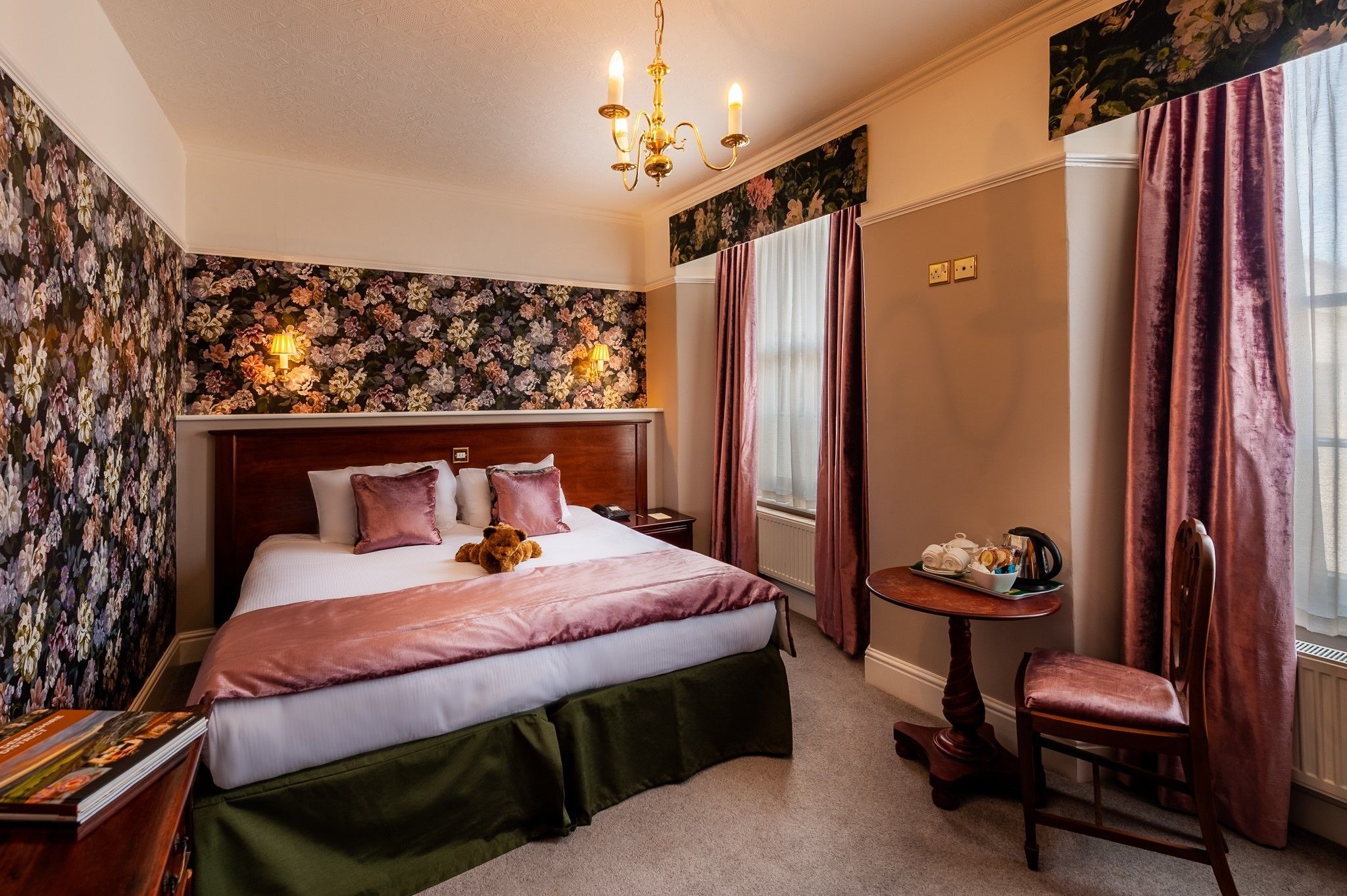 Een slaapkamer met een groot bed, een houten hoofdbord, een klein tafeltje met stoel, en behang met een bloemenmotief.