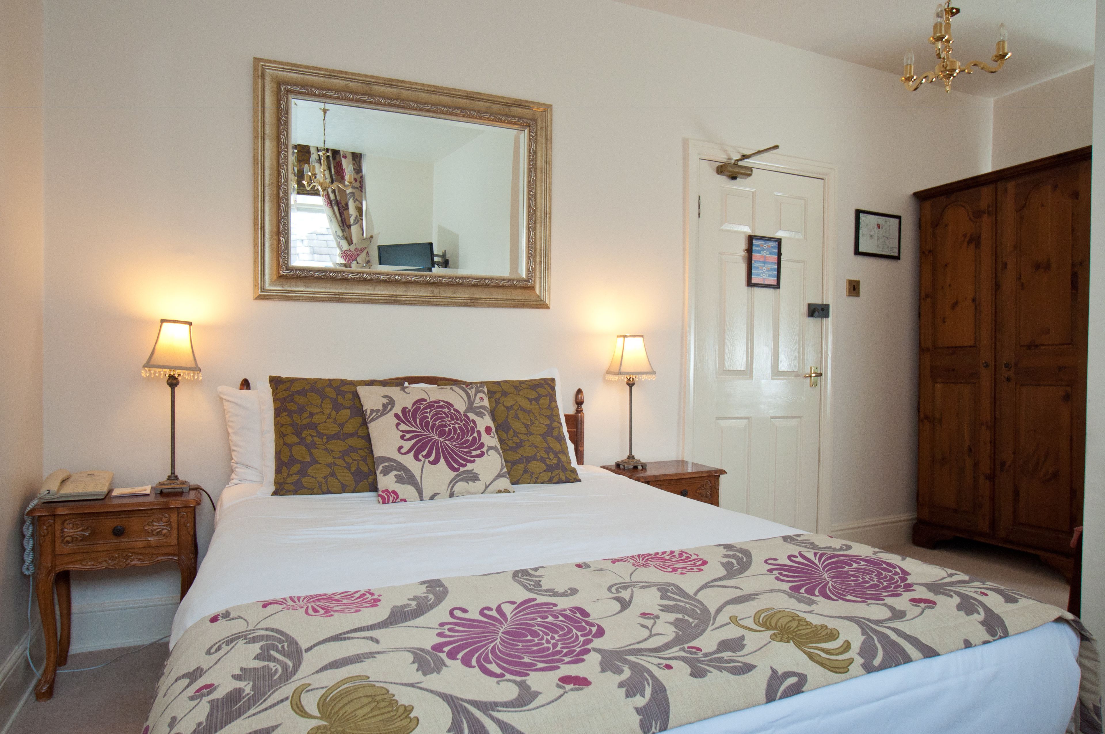 A neatly arranged bedroom with a large bed, two bedside tables with lamps, a wooden wardrobe, and a door with a mirror above it.