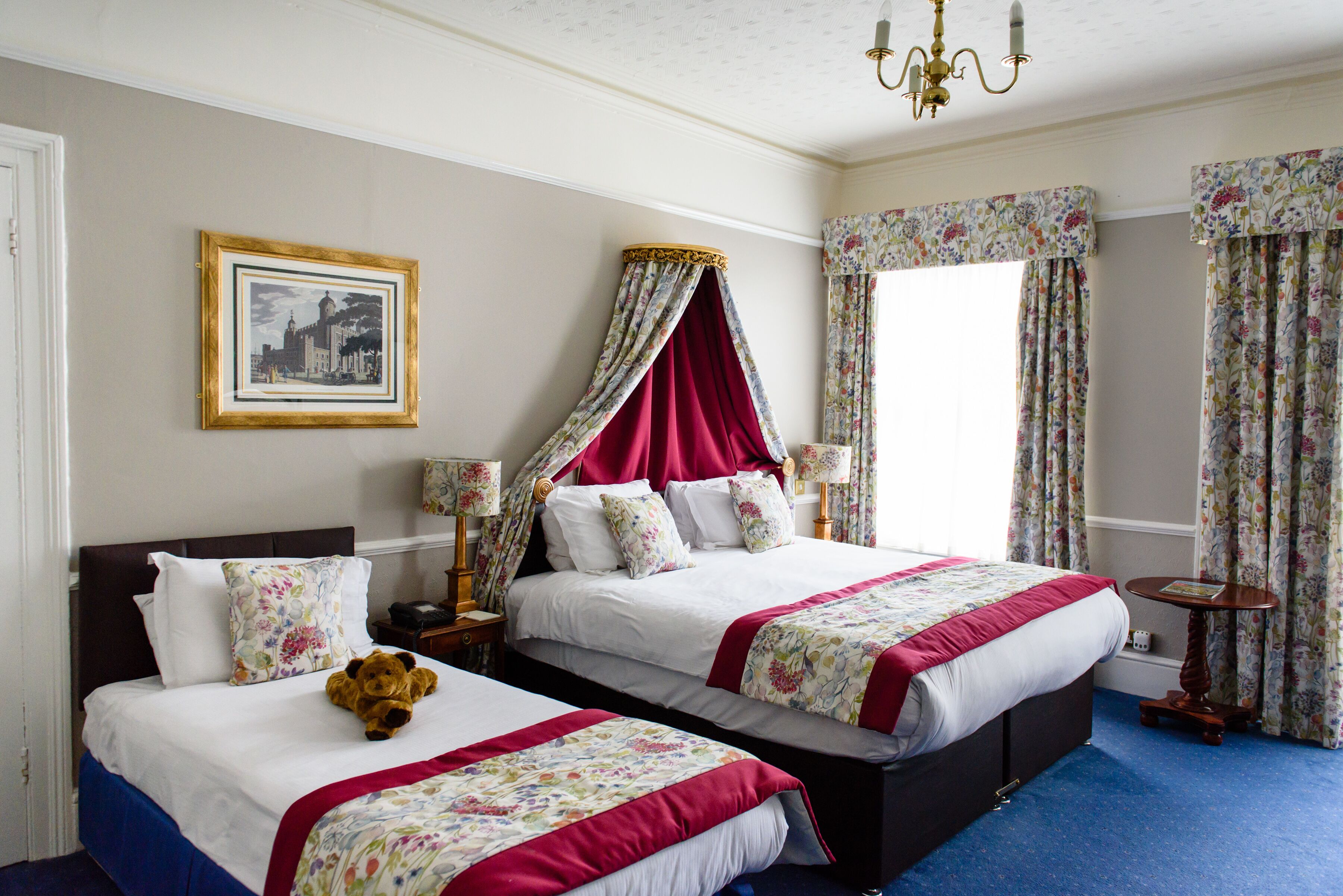 A double bed with floral bedding, a nightstand with a lamp, a teddy bear, and a framed picture on the wall.