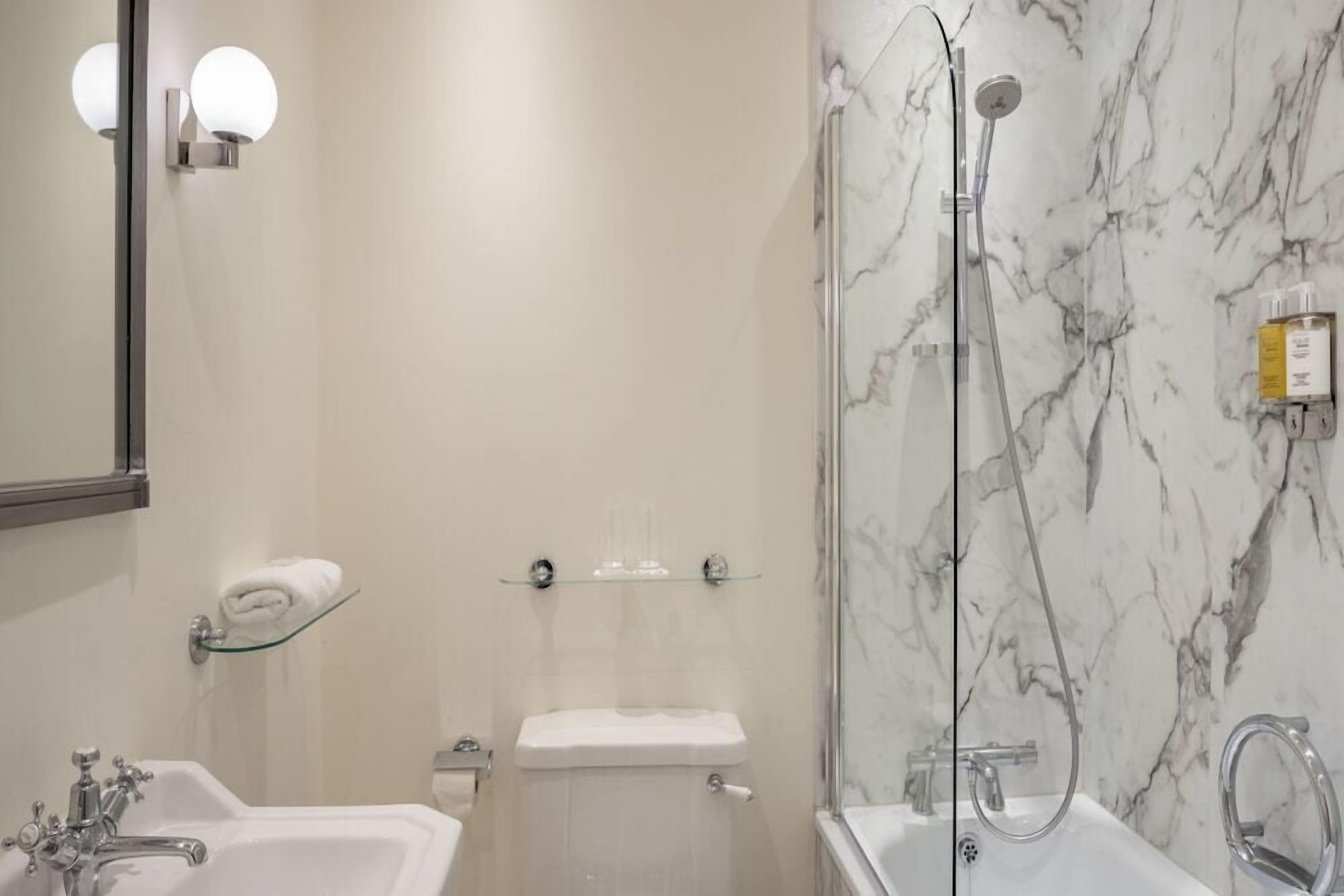 Un baño con pared de mármol, repisa de vidrio con artículos de aseo, lavamanos blanco y bañera.