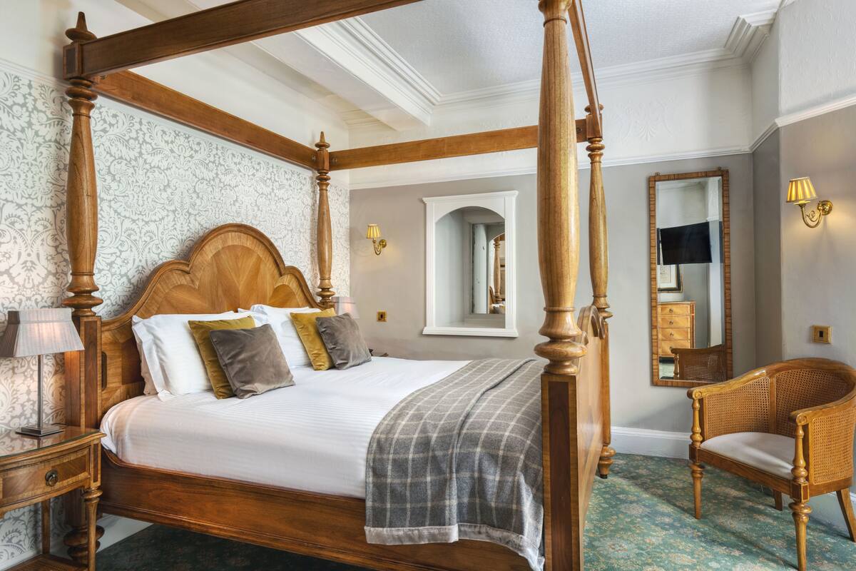 A four-poster bed with a wooden headboard, a bedside table with a lamp, a chair, and a mirror on the wall.
