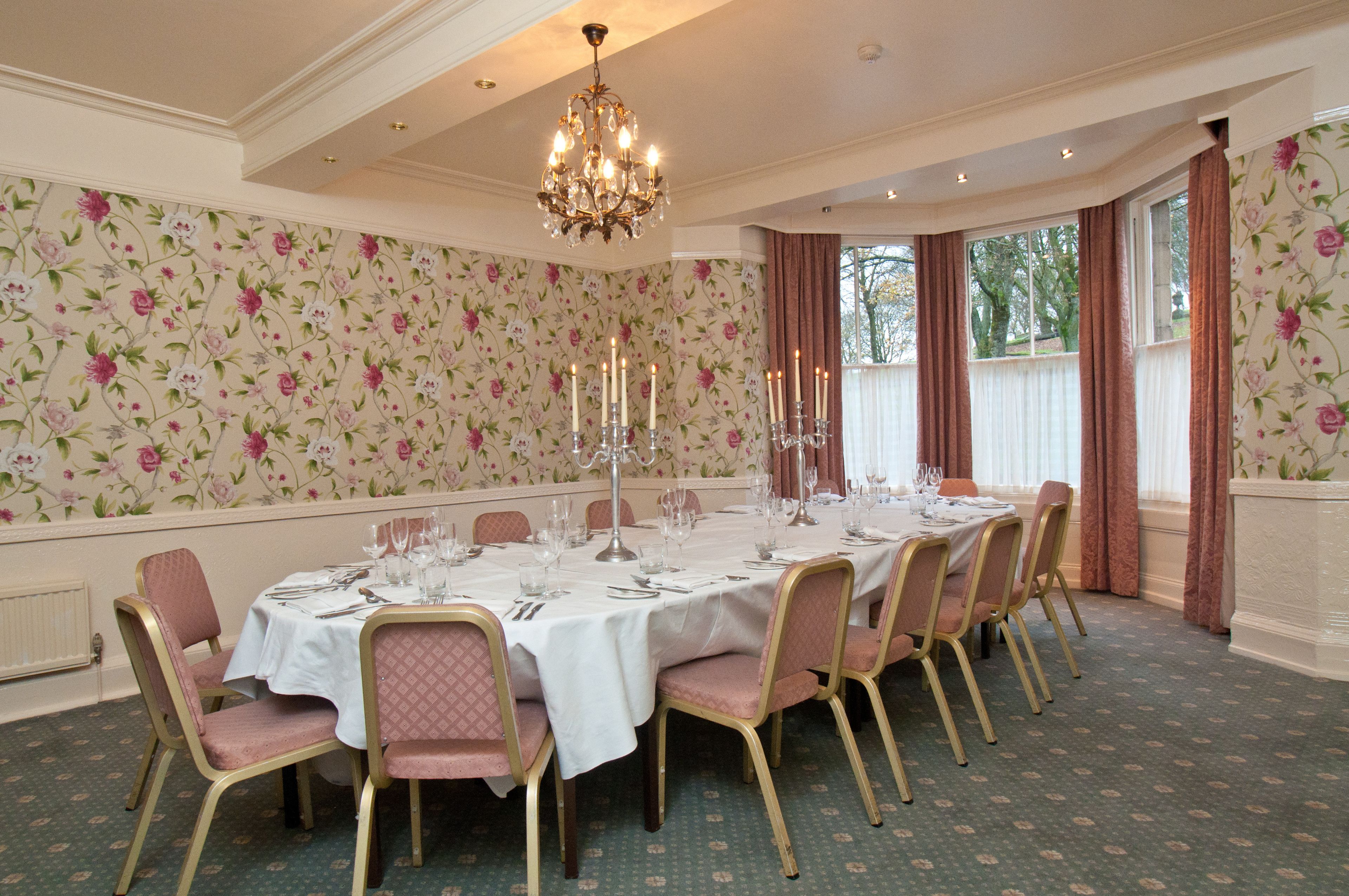 A formal dining room with a long table set for a meal, floral wallpaper, and a chandelier.