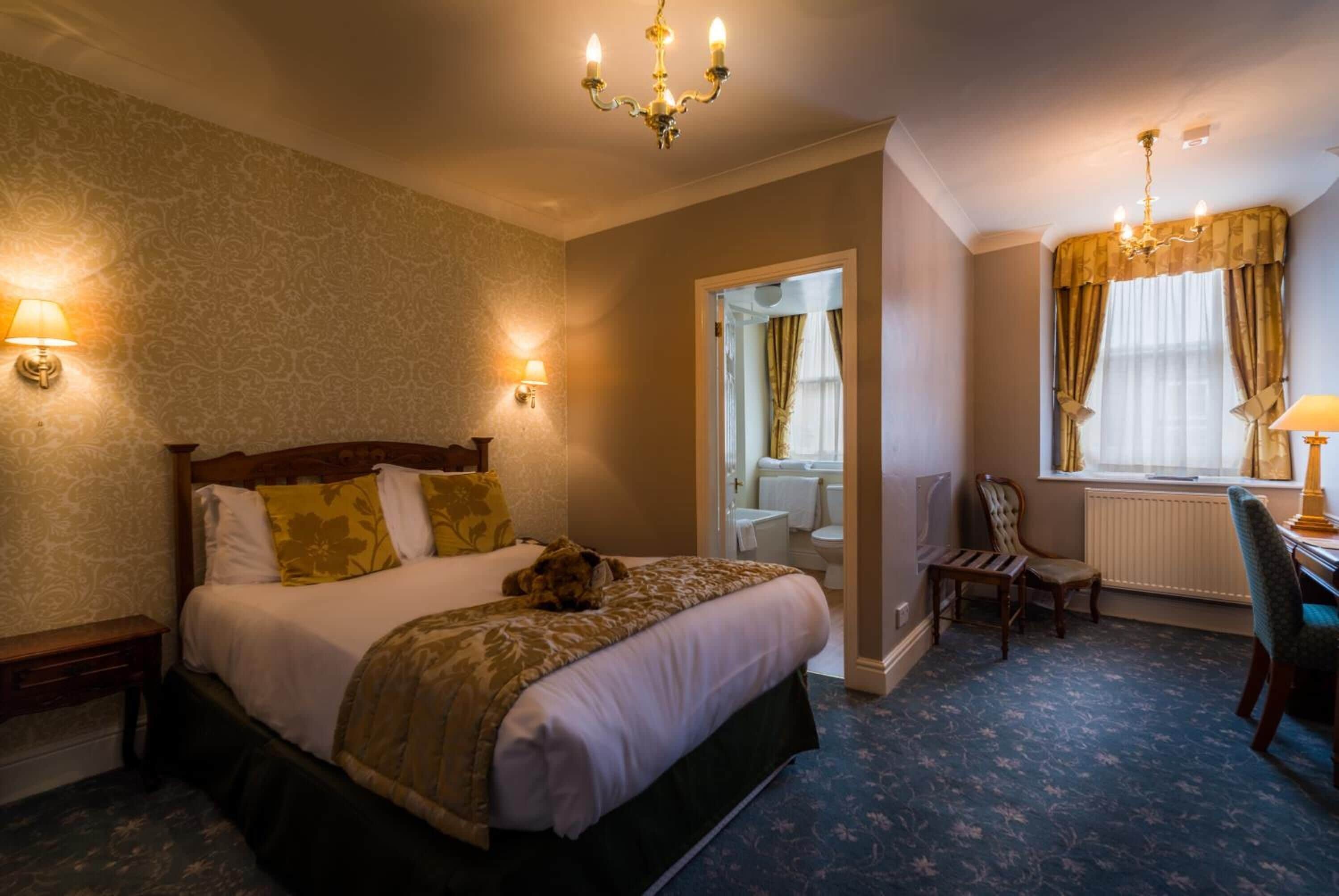 A hotel room with a large bed, two bedside lamps, a chandelier, a window with curtains, a chair, and a desk with a lamp.