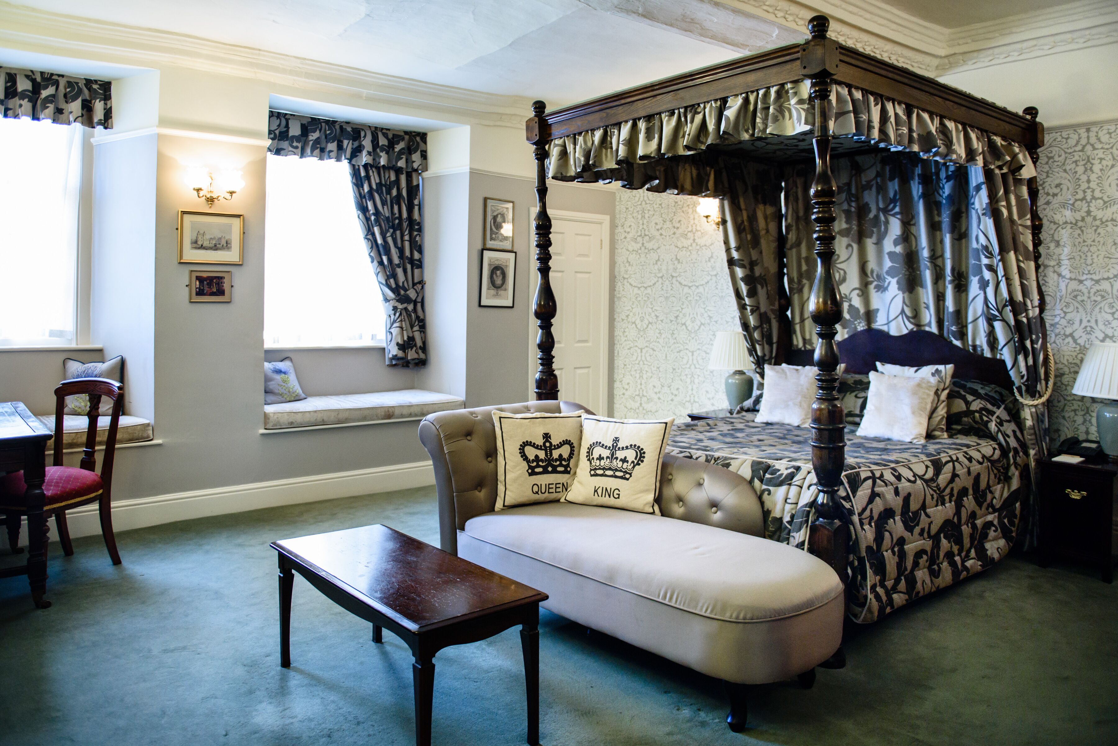A four-poster canopy bed with a tufted headboard, a chaise lounge, and a small table in a room with patterned wallpaper and a window with curtains.
