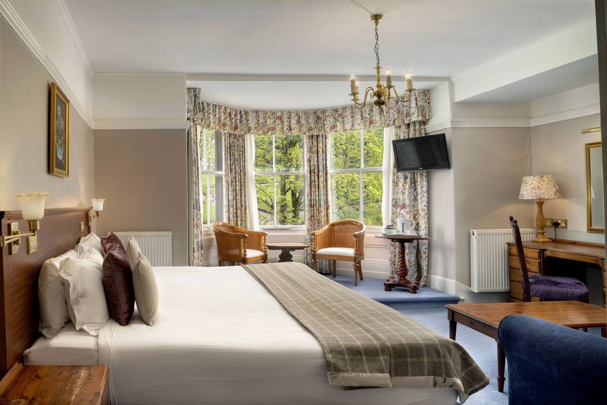 A hotel room with a large bed, two chairs, a small table, a TV, and a window with curtains.
