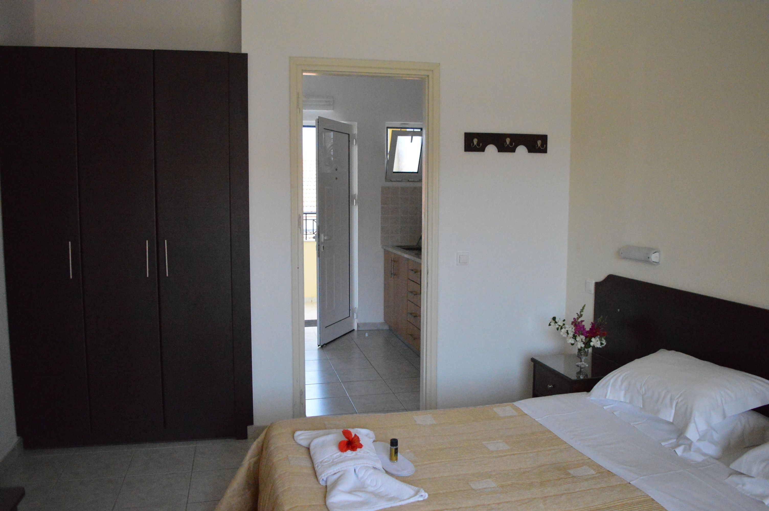 Habitación de hotel con cama, mesita de noche con un florero, armario y vistas al baño y la cocina a través de una puerta abierta.