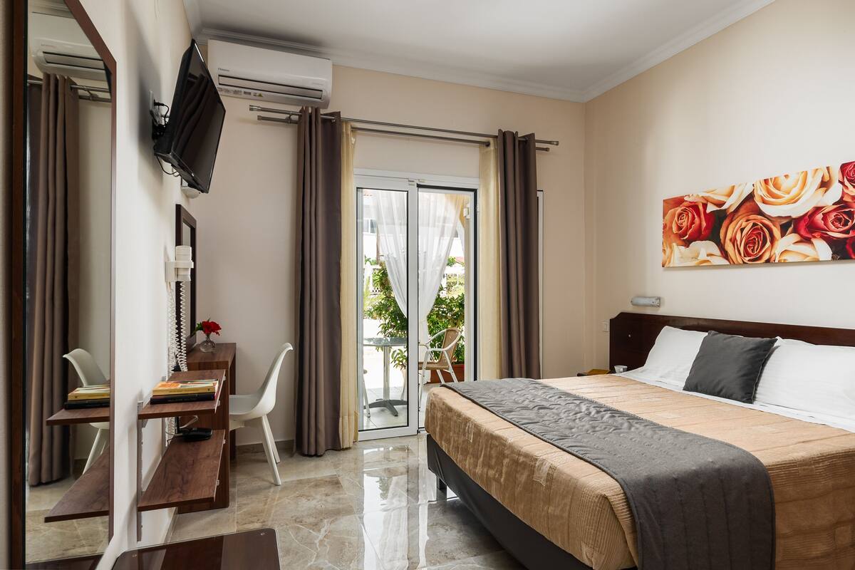A hotel room with a bed, a desk with a chair, a TV mounted on the wall, and a sliding glass door leading to an outdoor area.