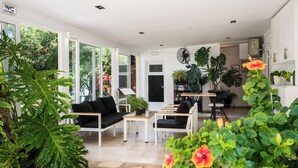 A spacious living room with a black sofa, a coffee table, and a dining area with a small table and chairs. There are large windows providing natural light and a view of greenery outside. - Konstantina Apartments (Corfu)