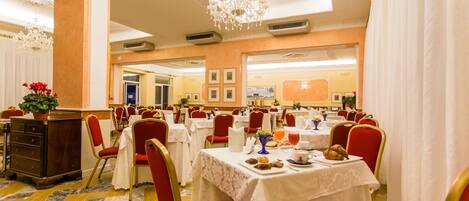 A hotel dining area with multiple tables set for breakfast, red chairs, and a chandelier.