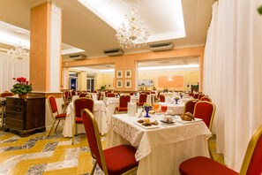 Une salle à manger d’hôtel avec plusieurs tables dressées pour le petit-déjeuner, des chaises rouges et un lustre.