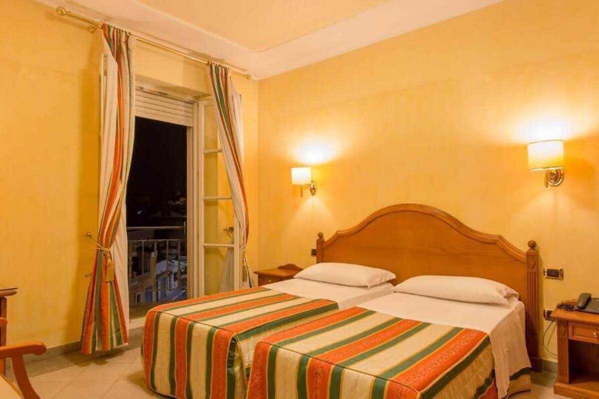 Een hotelkamer met een bed, nachtkastjes, een stoel, een raam met gordijnen en wandlampen.