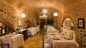 Un restaurante con paredes de ladrillo y techos abovedados, mesas dispuestas con manteles blancos y sillas traslúcidas, y una araña colgando del techo.